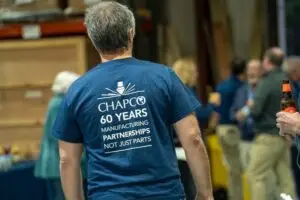 Chapco Sheet Metal Fabrication | A man with gray hair wears a blue T-shirt that reads, CHAPCO 60 YEARS MANUFACTURING PARTNERSHIPS NOT JUST PARTS, highlighting Chapco's contribution. He stands indoors while several people are gathered and talking in the background. | Deep River, CT