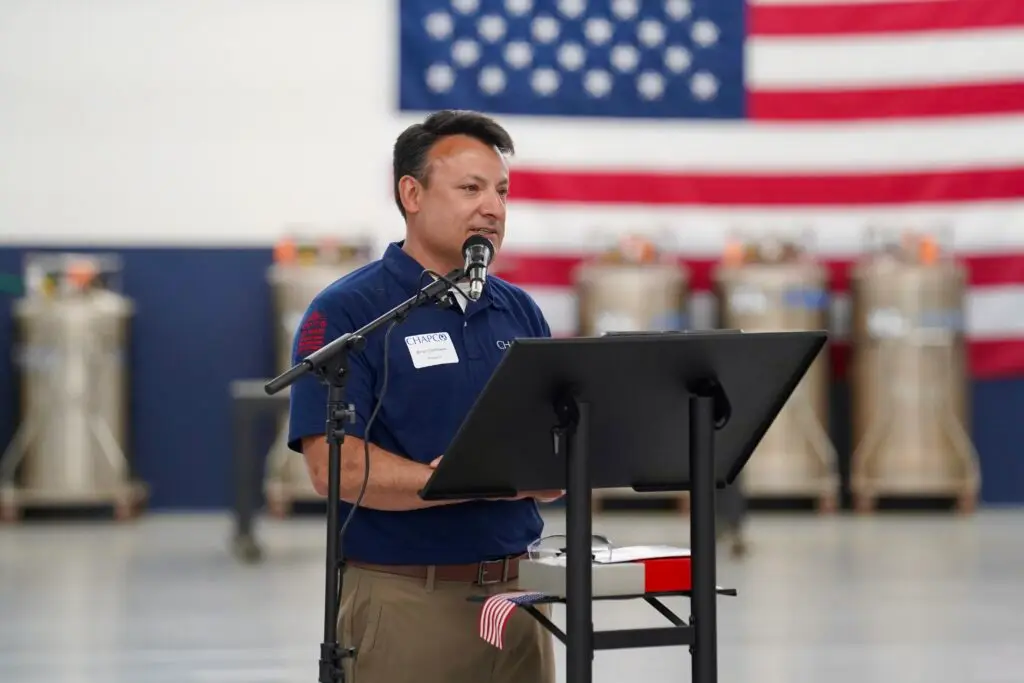 Man in a blue polo shirt speaks into a microphone at a podium, with a large American flag in the background.