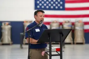 Man in a blue polo shirt speaks into a microphone at a podium, with a large American flag in the background.